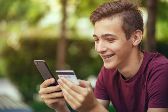 Teenage boy with a credit card and phone makes purchasing outdoors. Happy young man is using smart phone and bank card for online shopping. Handsome smiling white guy holds bank  card and cell phone.